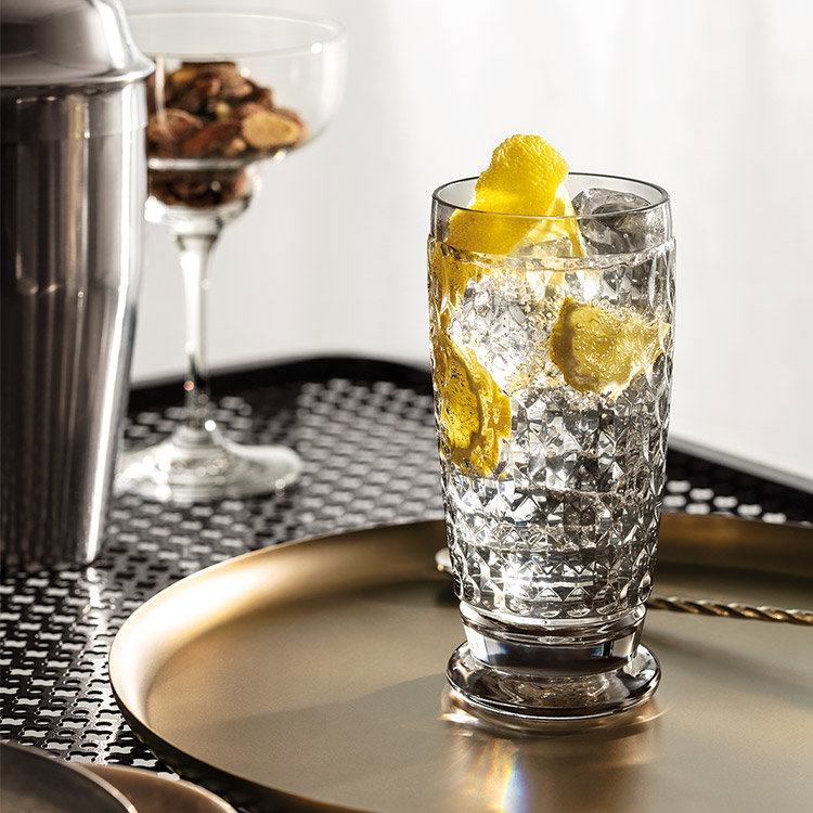 Cocktail mit Eis und Zitronenzeste im Glas auf Tablett, Barzubehör im Hintergrund.