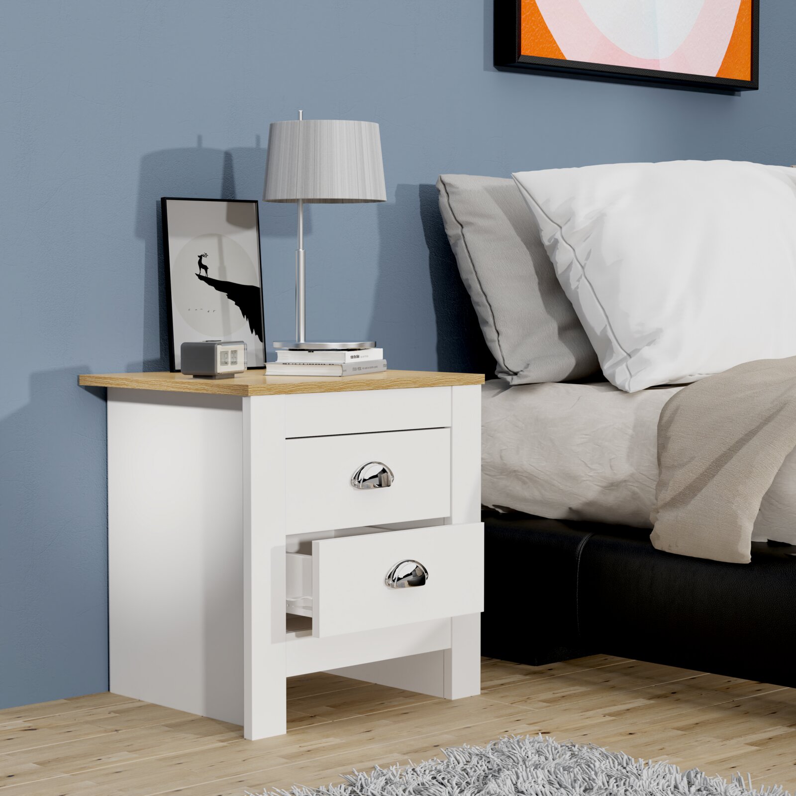 Weißer Nachttisch mit zwei Schubladen und Eichenholz-Optik-Oberplatte, eine Schublade halb geöffnet darauf eine silberne Tischlampe, Bücher und ein gerahmtes Bild neben einem Bett – Perspektive: frontale 3/4-Ansicht von links.