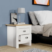 Weißer Nachttisch mit zwei Schubladen und Eichenholz-Optik-Oberplatte, eine Schublade halb geöffnet; darauf eine silberne Tischlampe, Bücher und ein gerahmtes Bild neben einem Bett – Perspektive: frontale 3/4-Ansicht von links.