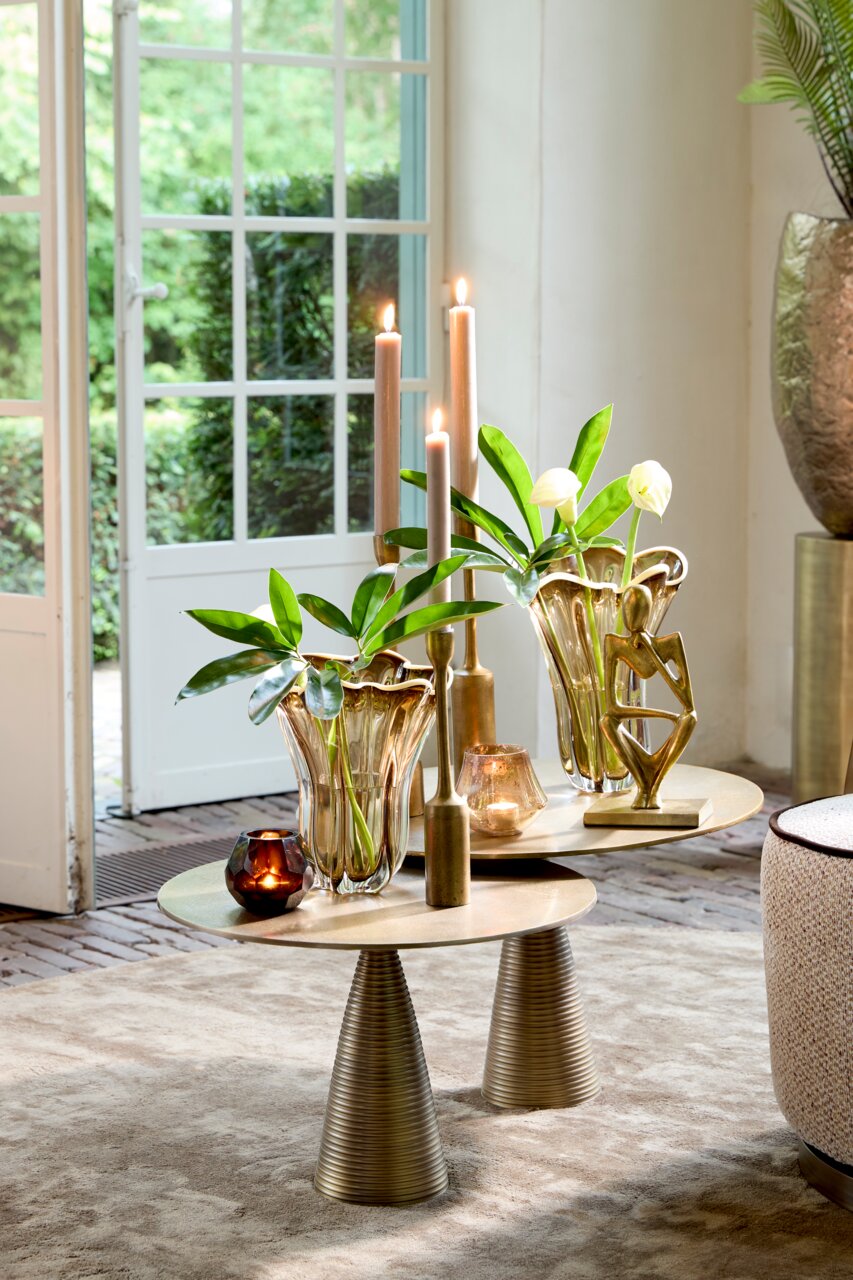 Elegante Vase mit wellenförmigem Rand, gefüllt mit grünen Blättern, auf einem stilvollen Tisch in einem hellen Raum, seitliche Perspektive.