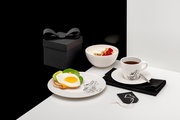 Schwarz-weißes Frühstücks-Setting in schräger Seitenansicht: weißes Geschirr mit Cartoon-Motiv, Tasse Kaffee auf Untertasse mit Löffel, Teller mit Toast, Spiegelei, Avocado und Käse, Schale Joghurt mit Nüssen und Himbeeren sowie schwarze Geschenkbox mit S