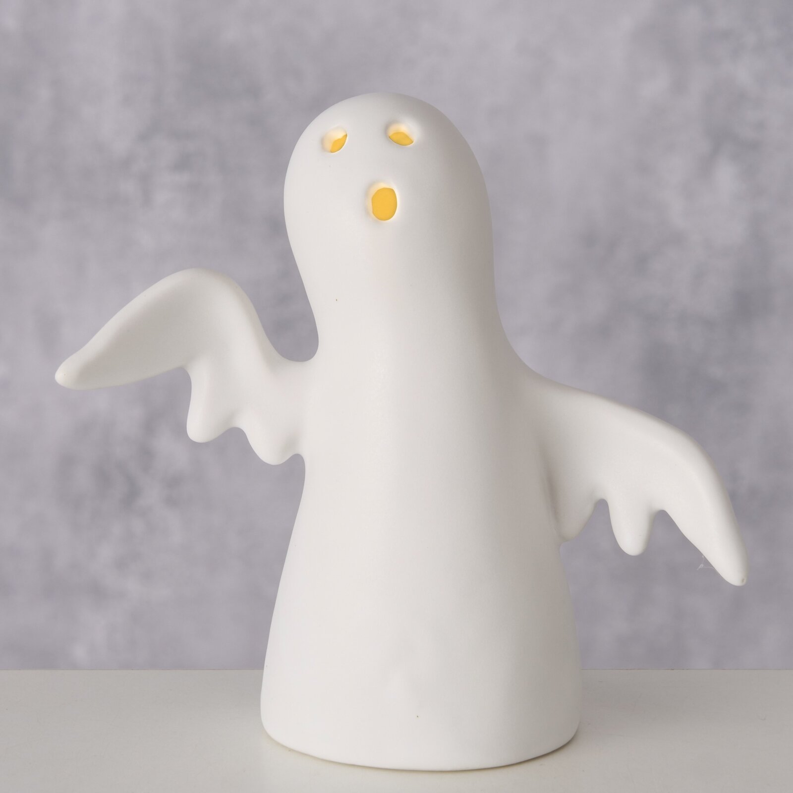 Keramisch beeldje van een spook met uitgespreide vleugels, van voren gezien, met stralende ogen en mond.