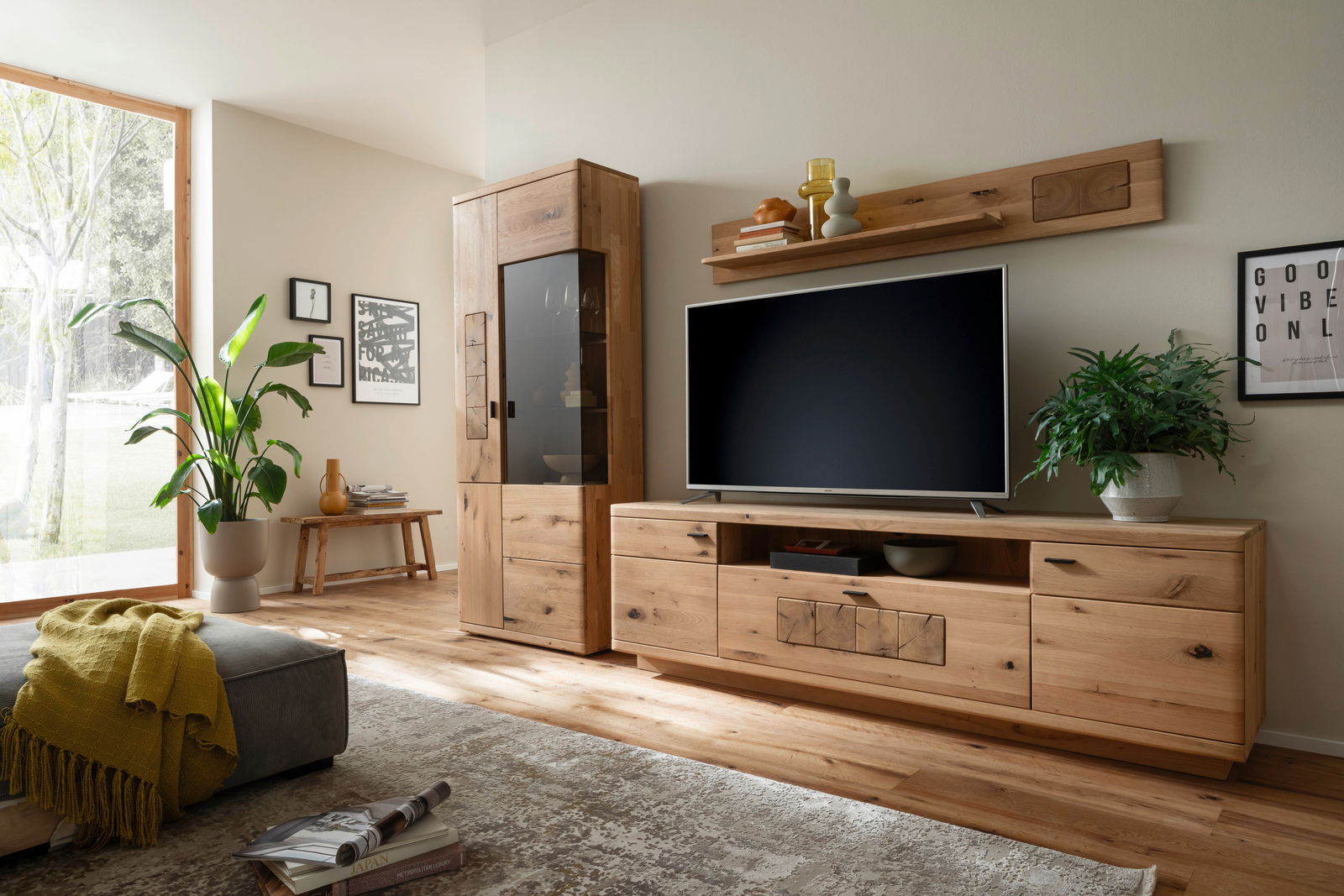 Moderne Wandkombination aus Holz in einem Wohnzimmer, bestehend aus einem hohen Schrank mit Glastür und einem breiten TV-Board mit Stauraum. Perspektive von schräg vorne.
