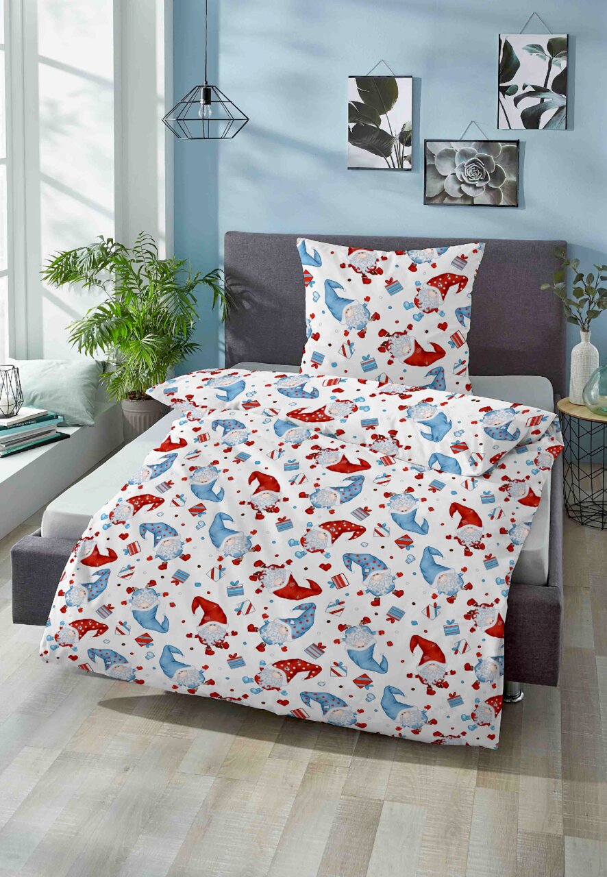 Biber Bettwäsche 100x135cm mit buntem Muster aus roten und blauen Mützen und Geschenken, auf einem Bett in einem hellen Raum mit Pflanzen und Wanddekorationen, aus der Perspektive von vorne fotografiert.