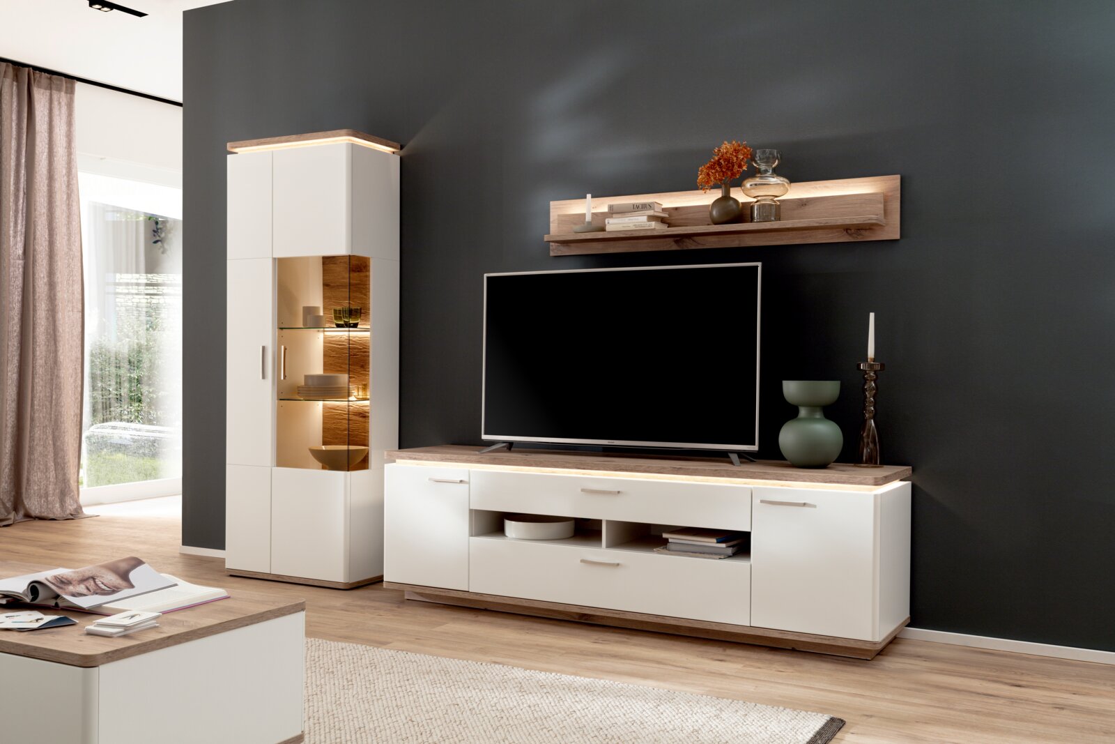 Moderne Wohnzimmer-Wohnwand in Weiß und Holzoptik mit TV-Lowboard, beleuchtetem Vitrinenschrank und Wandregal vor dunkler Wand, Ansicht schräg von vorn