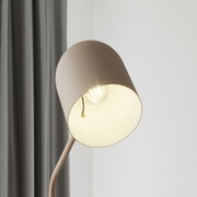 Close-up van een moderne staande lamp met een eenvoudige, ronde lampenkap in beige, die een gloeilamp omhult. De foto toont de lamp van opzij, met een grijs gordijn op de achtergrond.