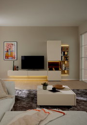 Frontale Ansicht einer modernen, beigefarbenen Wohnwand mit schwebendem TV-Lowboard, hohen Schrankelementen, beleuchteter Vitrine und offenen Holzregalen in einem stilvollen Wohnzimmer.