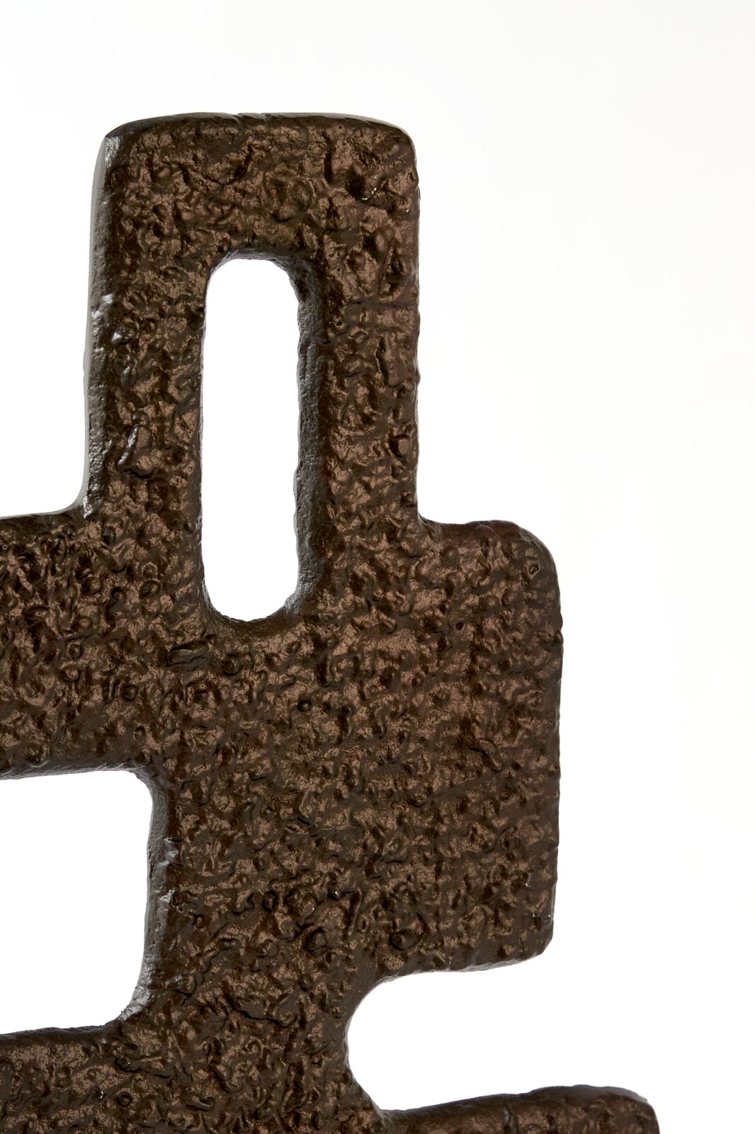 Close-up in vooraanzicht van een abstract donkerbruin metalen object met geometrische openingen en een robuust, organisch reliëf.