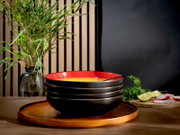 Schwarze Suppenschalen mit roter Innenseite, frontal aus leichter Obersicht auf einem runden Holztablett arrangiert, daneben Limetten, Radieschen und frische Kräuter.