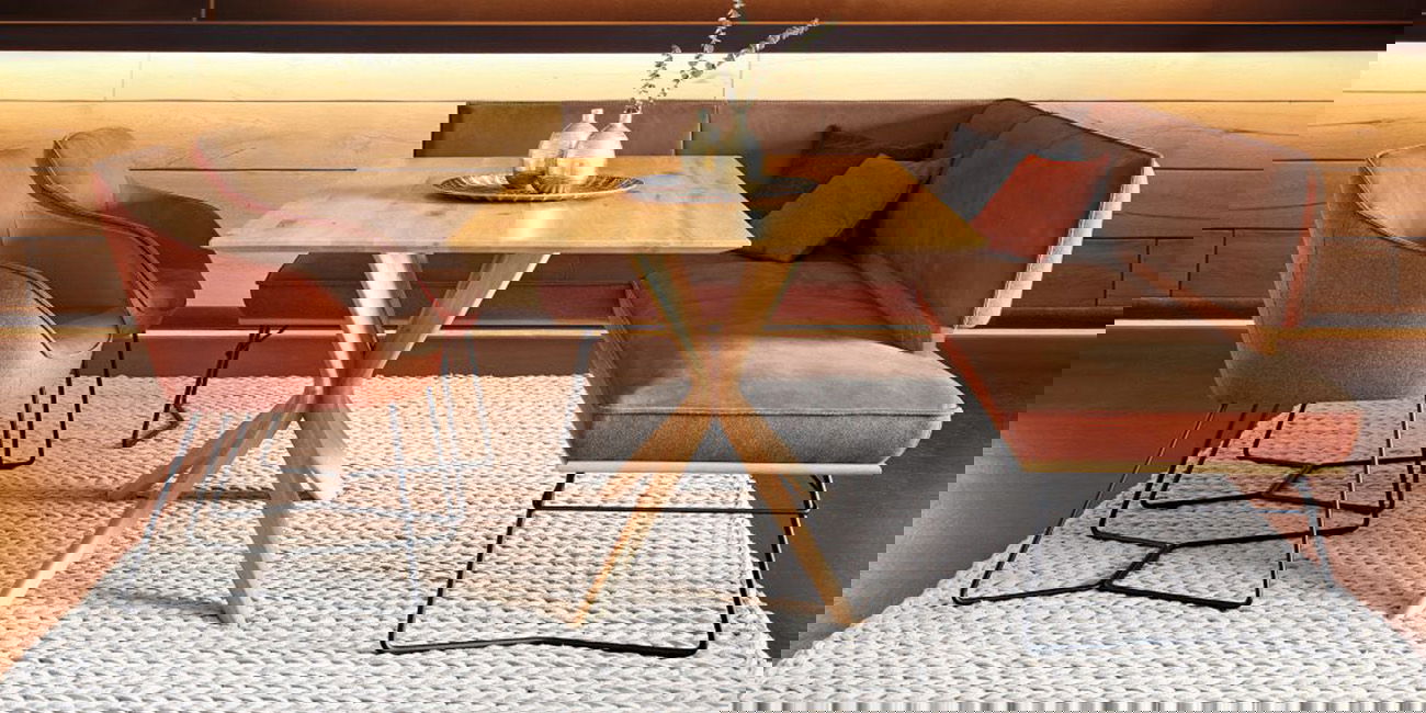 Auf dem Bild sieht man einen Holztisch mit Stühlen und einer Eckbank. Die Möbel haben weiche Polster in Braun und Orange. Der Teppich darunter ist hell. Alles sieht gemütlich und modern aus.