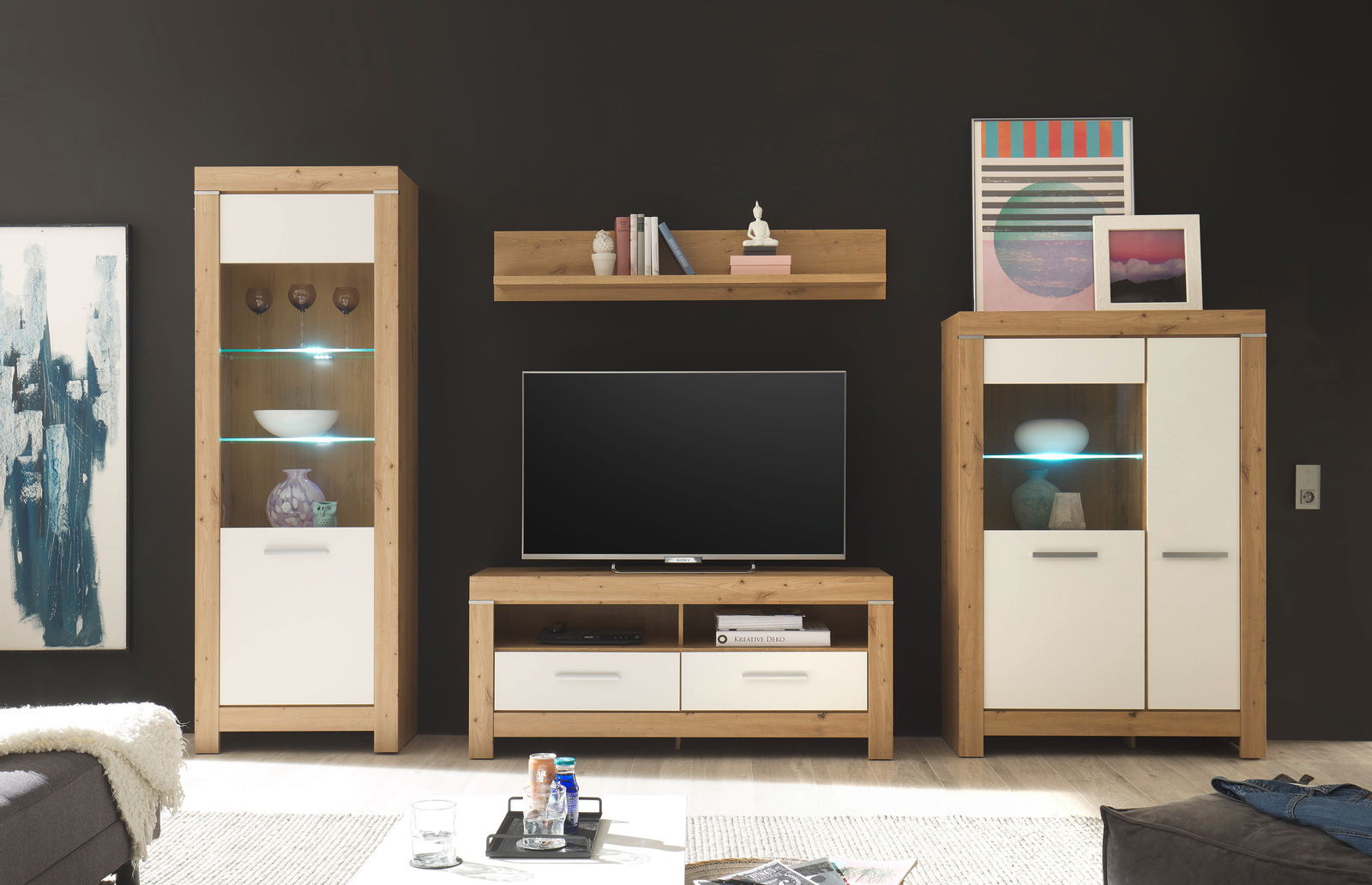 Moderne Wohnwand in Holzoptik mit weißem Akzent, bestehend aus zwei Vitrinen, einem TV-Board und einem Wandregal, frontal betrachtet.