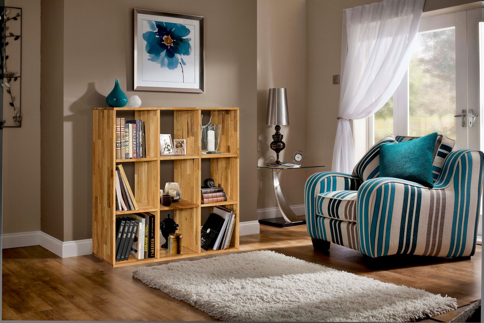 Holzregal mit mehreren Fächern, gefüllt mit Büchern und Dekorationen, in einem gemütlichen Wohnzimmer mit einem gestreiften Sessel und einem großen Fenster. Perspektive von vorne.