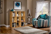 Holzregal mit mehreren Fächern, gefüllt mit Büchern und Dekorationen, in einem gemütlichen Wohnzimmer mit einem gestreiften Sessel und einem großen Fenster. Perspektive von vorne.