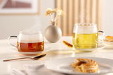 Zwei Teetassen aus Glas, gefüllt mit dampfendem Tee, auf einem Tisch mit Gebäck und einem Löffel, aus seitlicher Perspektive fotografiert.