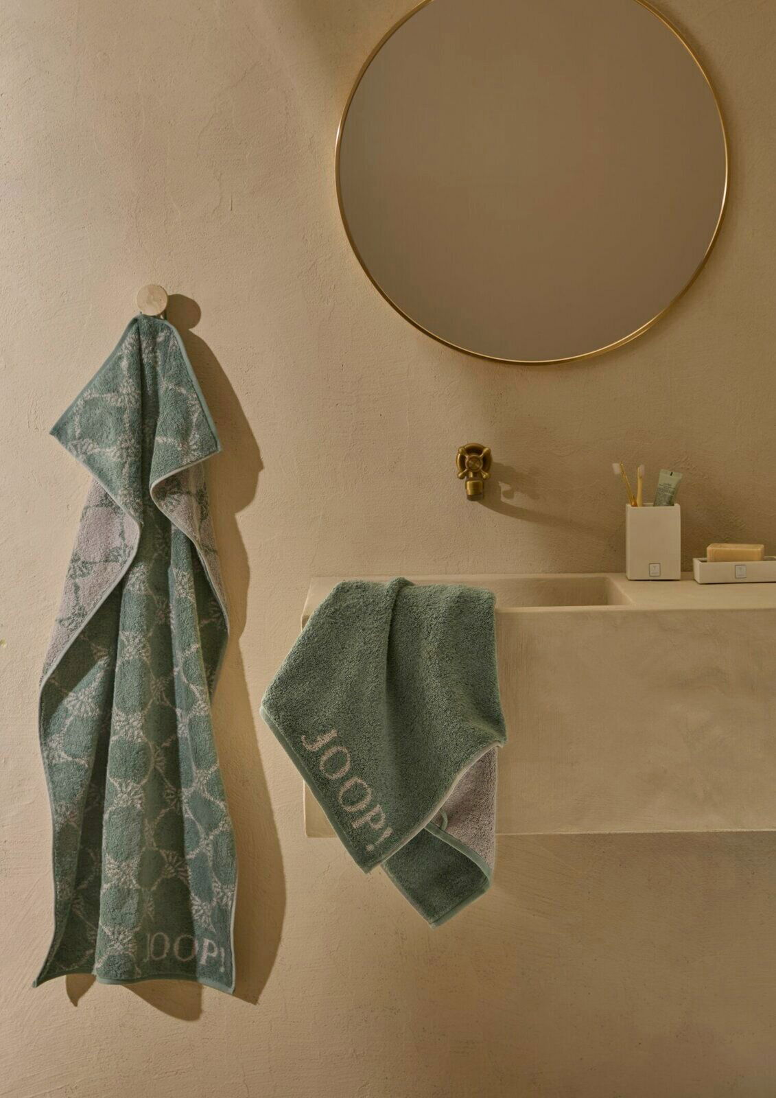 Gästetuch in einem eleganten Badezimmer, aufgehängt an einem Haken und über einem Waschbecken drapiert, mit einem runden Spiegel im Hintergrund. Perspektive von vorne.