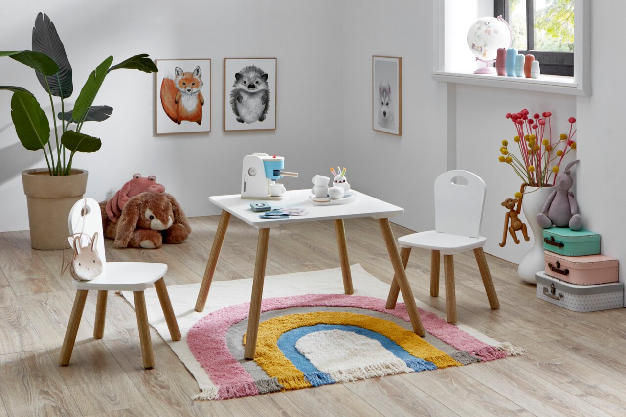 Kinderzimmer-Szene mit weißem Kindertisch und zwei passenden Stühlen mit Holzbeinen auf buntem Regenbogen-Teppich, fotografiert in leichter Schrägansicht von vorn.