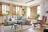 Gemütliche Wohnzimmer-Ecke mit einer großen, hellgrauen Rundecke, dekoriert mit Kissen und einer Decke. Im Vordergrund ein moderner Couchtisch, im Hintergrund große Fenster mit Holzjalousien. Perspektive: Frontalansicht auf die Rundecke.
