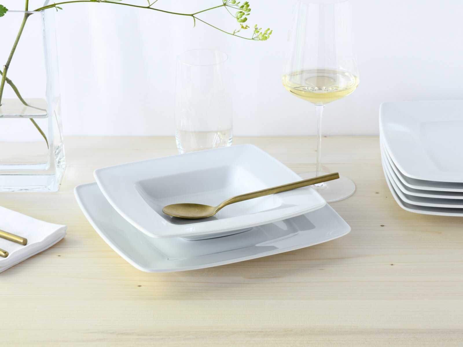 Schlichtes weißes quadratisches Geschirr-Set mit tiefem Teller/Schale und flachem Teller, darauf ein goldfarbener Löffel; seitliche Perspektive auf einem hellen Holztisch mit Weinglas und Wasserglas im Hintergrund