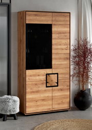 Hoher Wohnzimmerschrank in Eichenholz-Optik mit schwarzer Glasfront und dekorativem quadratischem Griffdetail, fotografiert in leichter Schrägansicht von vorn (Frontansicht).
