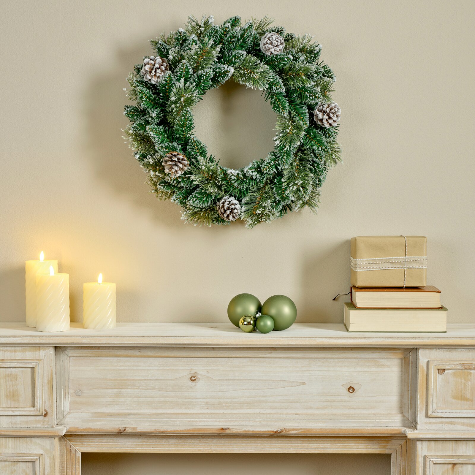 Vooraanzicht van een groene kerstkrans met besneeuwde dennentakken en dennenappels, sfeervol aan de muur boven een houten dressoir met brandende kaarsen, kerstballen en cadeaus