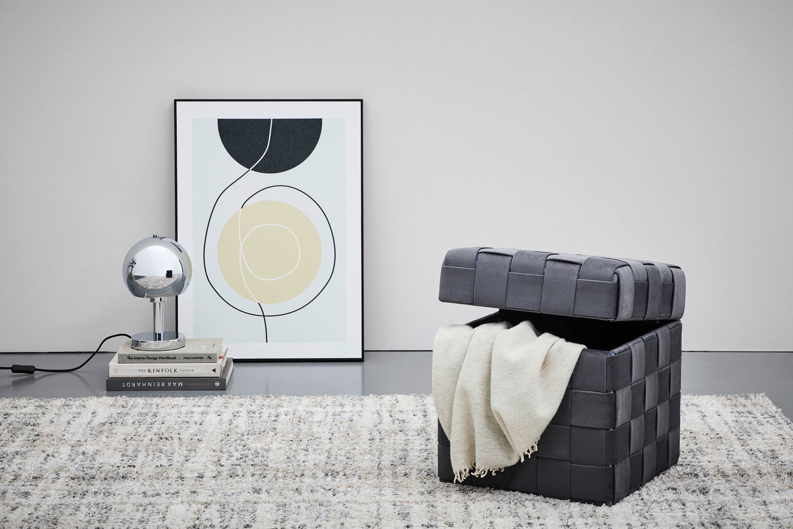 Grauer Hocker mit Stauraum, geöffnet, mit einer Decke darin, auf einem Teppich stehend, seitliche Perspektive.