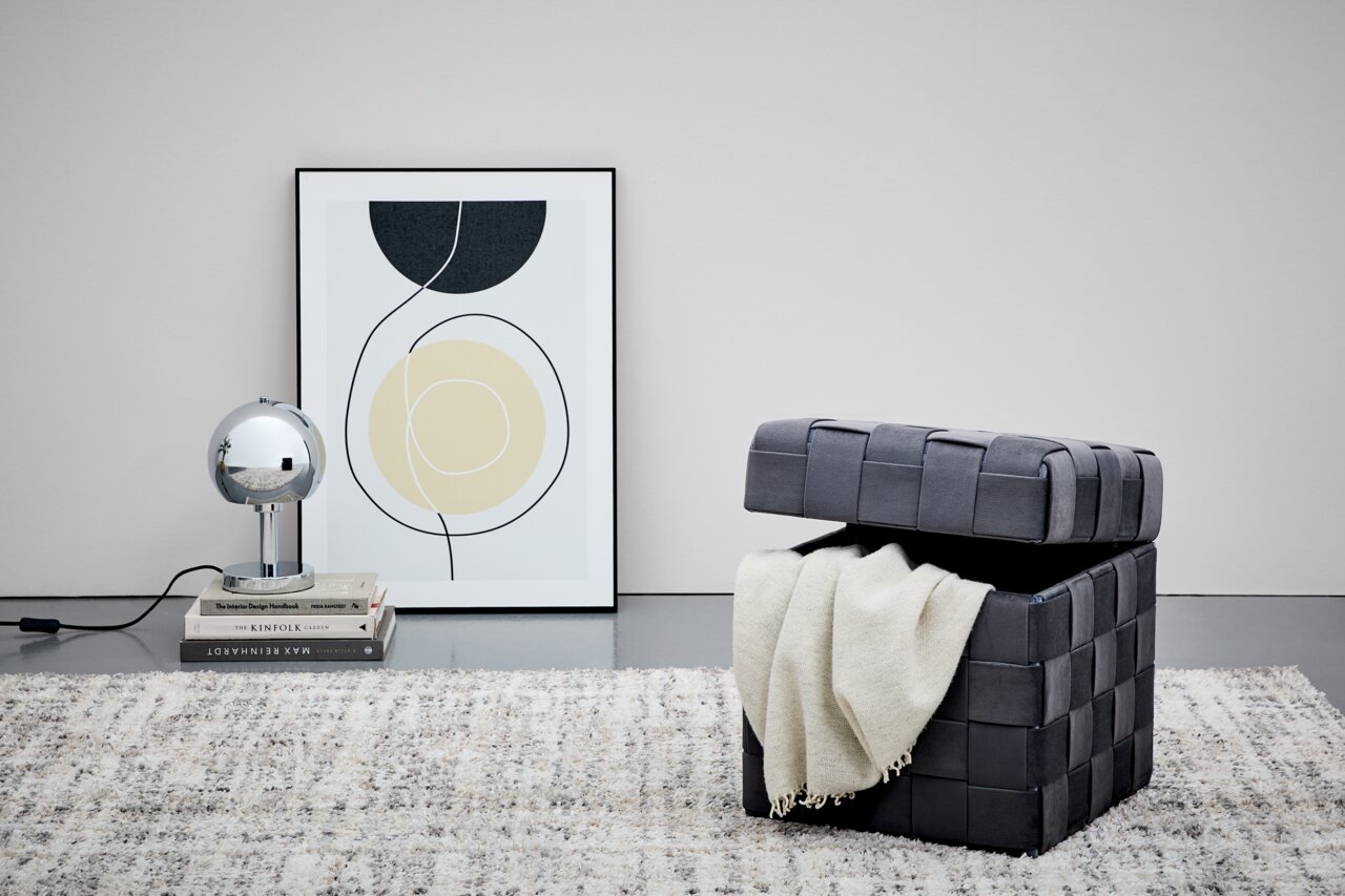 Grauer Hocker mit Stauraum, geöffnet, mit einer Decke darin, auf einem Teppich stehend, seitliche Perspektive.