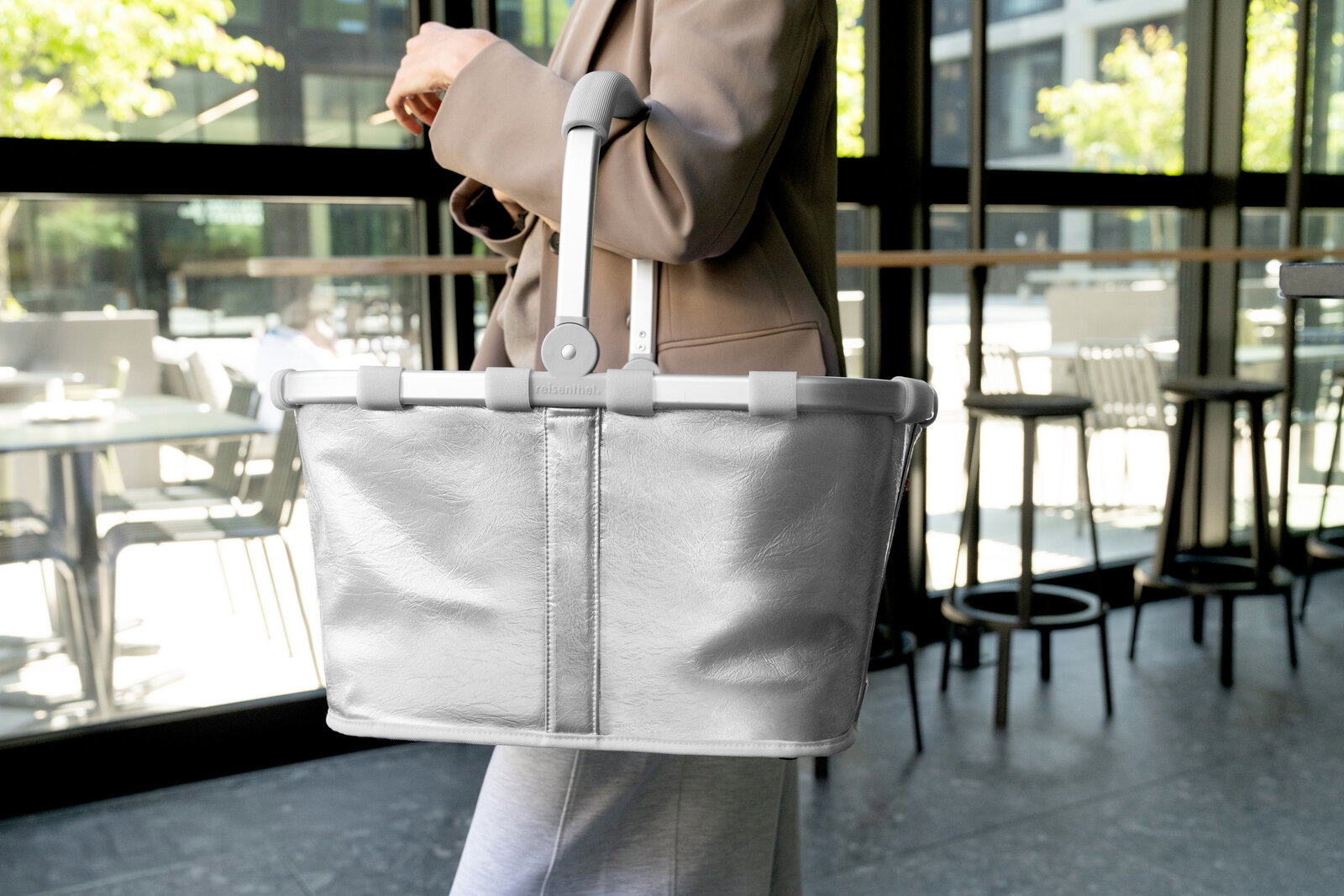 Silberner Einkaufskorb mit klappbaren grauen Griffen wird von einer Person in einem modernen Café getragen, Seitenansicht auf das Produkt.