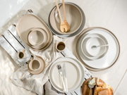 Draufsicht auf ein modernes, minimalistisches Geschirr-Set in Beige, Grau und Weiß mit Tellern, Schüsseln und Tassen, arrangiert auf weißer Holzfläche mit Leinentuch, Besteck und Holzsalatbesteck.