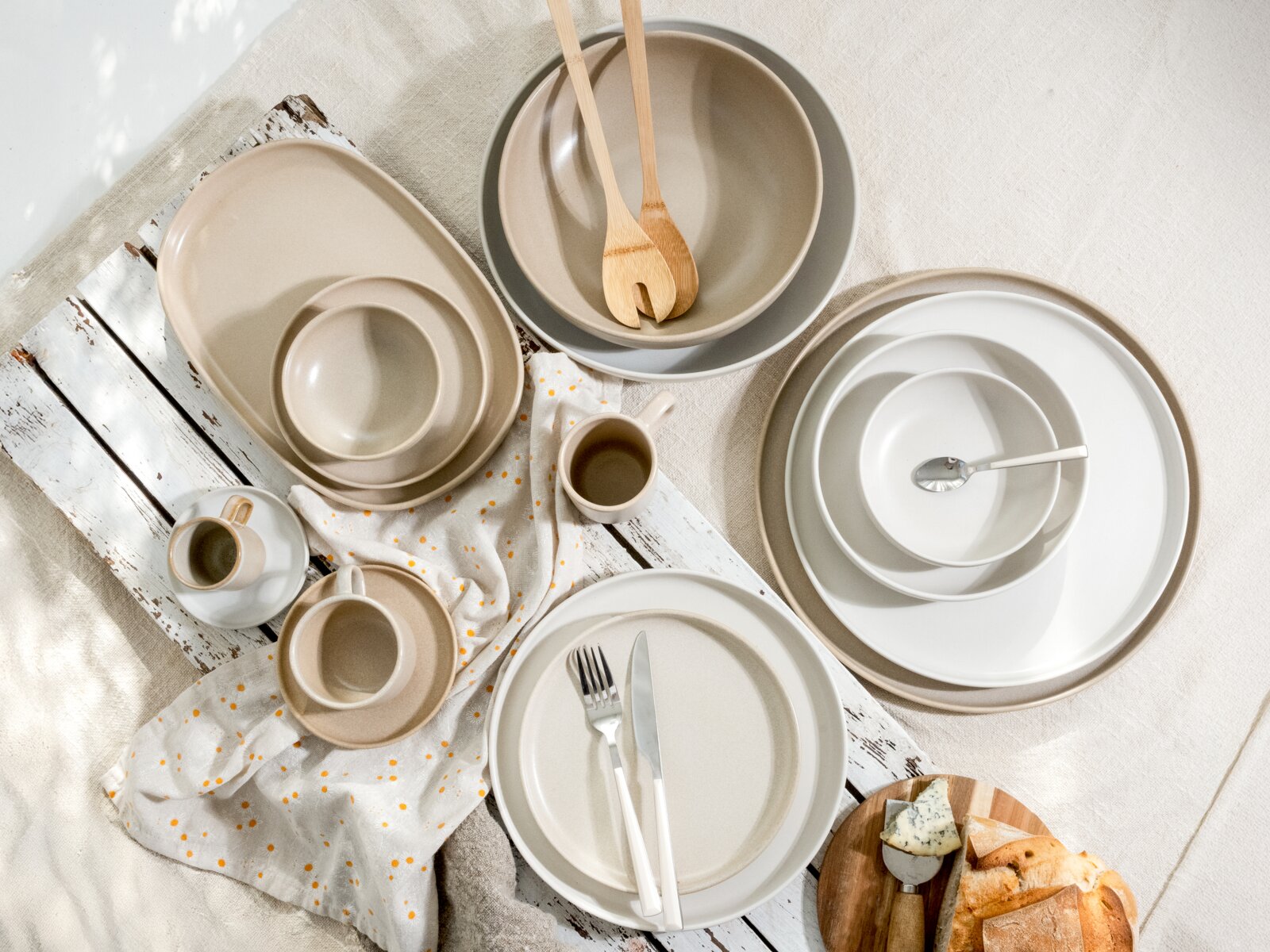 Draufsicht auf ein modernes, minimalistisches Geschirr-Set in Beige, Grau und Weiß mit Tellern, Schüsseln und Tassen, arrangiert auf weißer Holzfläche mit Leinentuch, Besteck und Holzsalatbesteck.