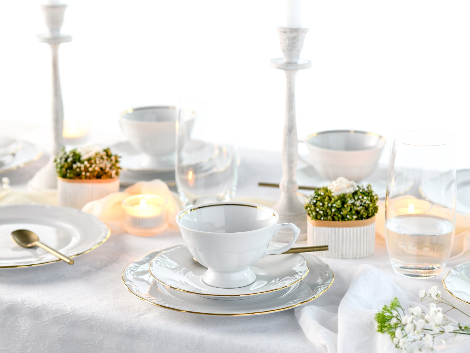 Elegant gedeckter Tisch mit weißem Porzellanservice mit goldfarbenem Rand (Tasse auf Untertasse und Teller), Kerzen und Gläsern, fotografiert aus leicht erhöhter, frontaler Perspektive.