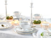 Elegant gedeckter Tisch mit weißem Porzellanservice mit goldfarbenem Rand (Tasse auf Untertasse und Teller), Kerzen und Gläsern, fotografiert aus leicht erhöhter, frontaler Perspektive.