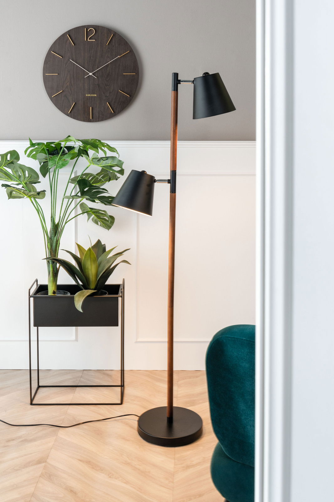 Modernes Wohnzimmer mit einer Stehlampe, einem Pflanzgefäß-Set mit grünen Pflanzen und einer Wanduhr. Perspektive von der Seite.