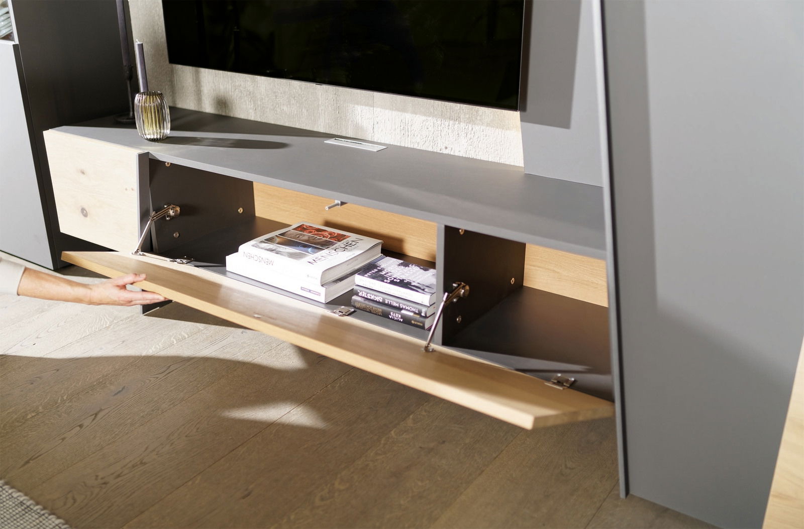 Nahaufnahme einer modernen Wohnwand mit grauer Oberfläche und Holzdetails, offene Klappe zeigt Stauraum mit Büchern.