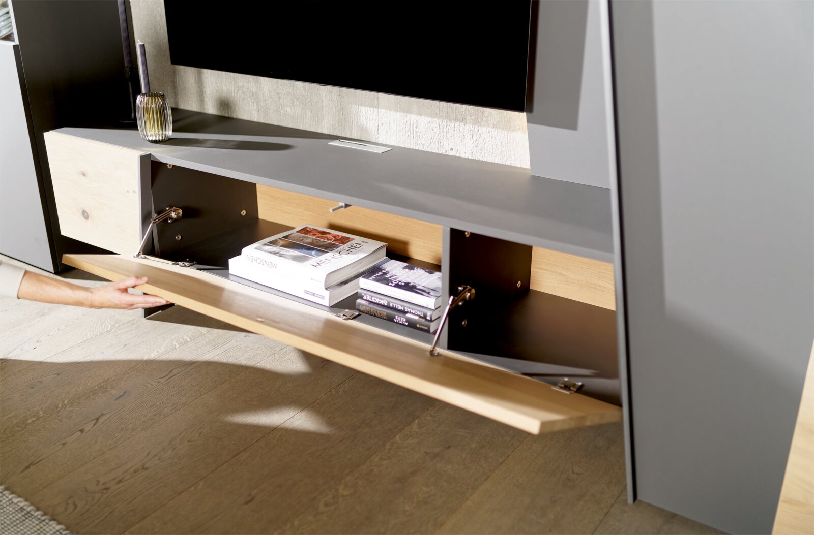 Nahaufnahme einer modernen Wohnwand mit grauer Oberfläche und Holzdetails, offene Klappe zeigt Stauraum mit Büchern.