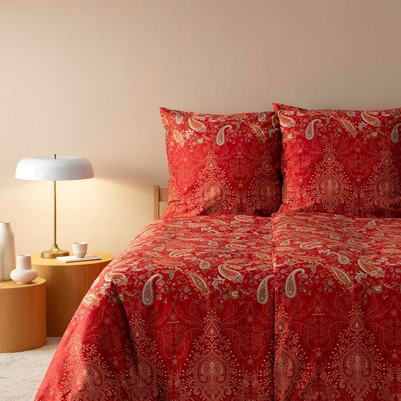 Rote Bettwäsche mit Paisley-Muster auf einem Doppelbett, seitliche Perspektive mit Nachttisch und Lampe.