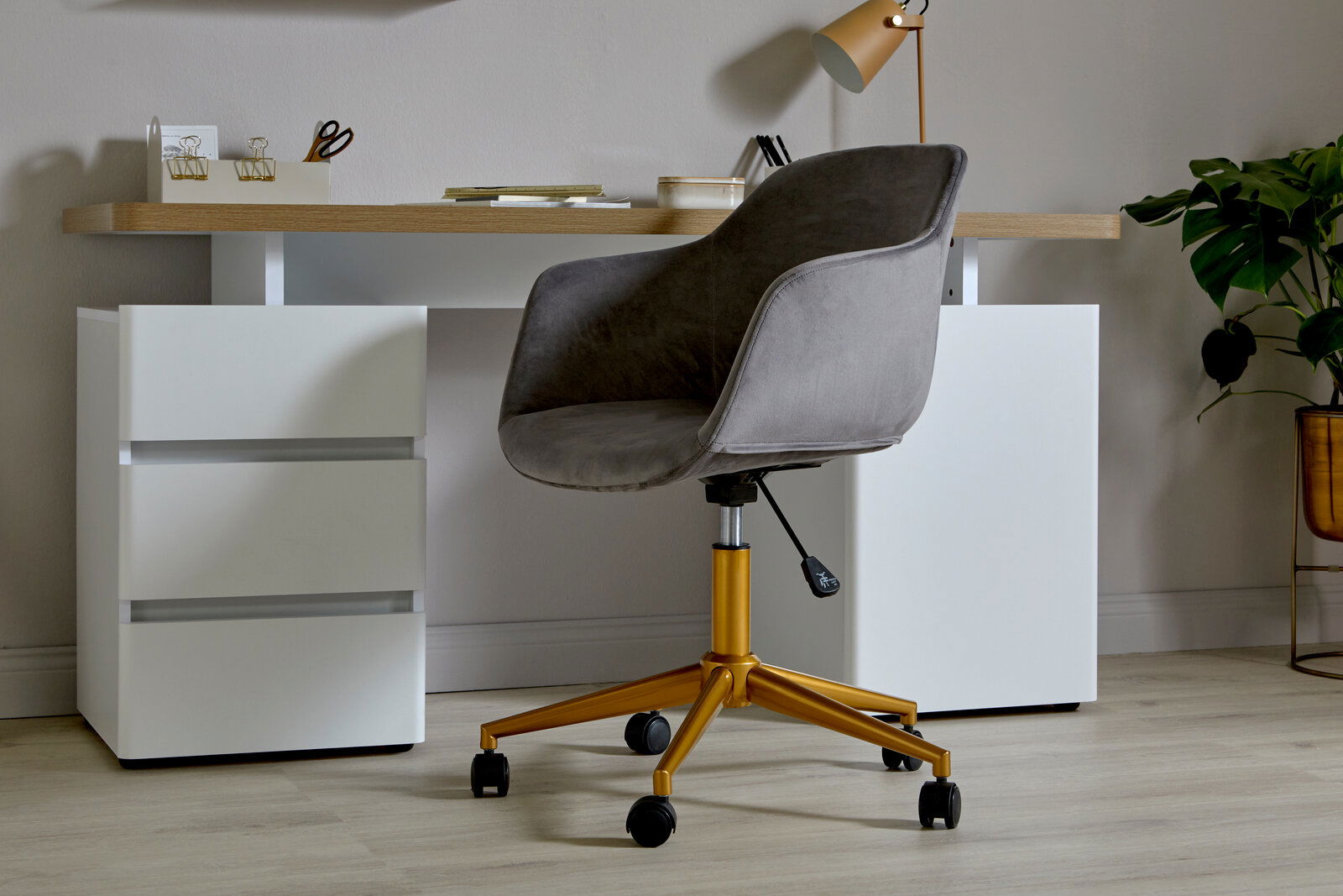 Grauer Bürostuhl mit goldenen Beinen und Rollen, seitliche Perspektive, vor einem modernen Schreibtisch mit weißen Schubladen.