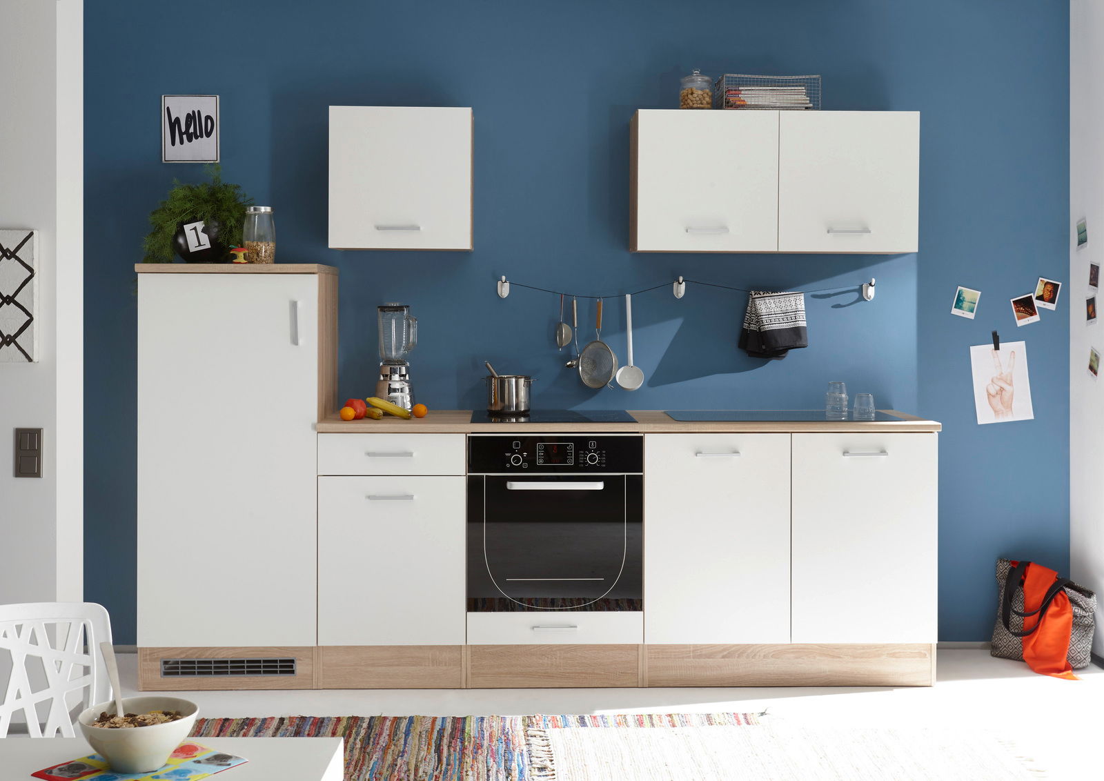 Moderne Küchenzeile mit weißen Schränken und Holzarbeitsplatte, frontal fotografiert vor einer blauen Wand.