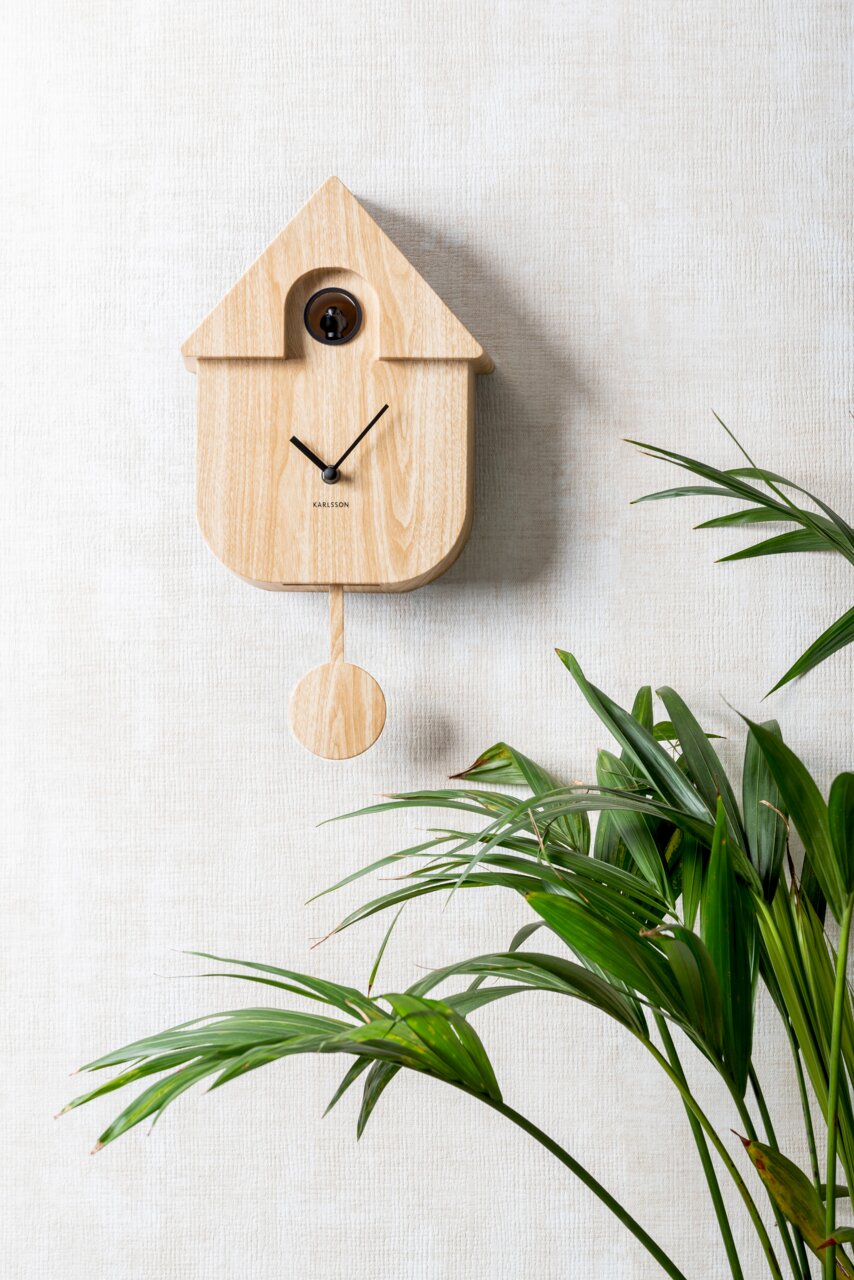 Moderne Wanduhr aus hellem Holz mit Pendel, frontal an einer strukturierten Wand neben einer grünen Pflanze.