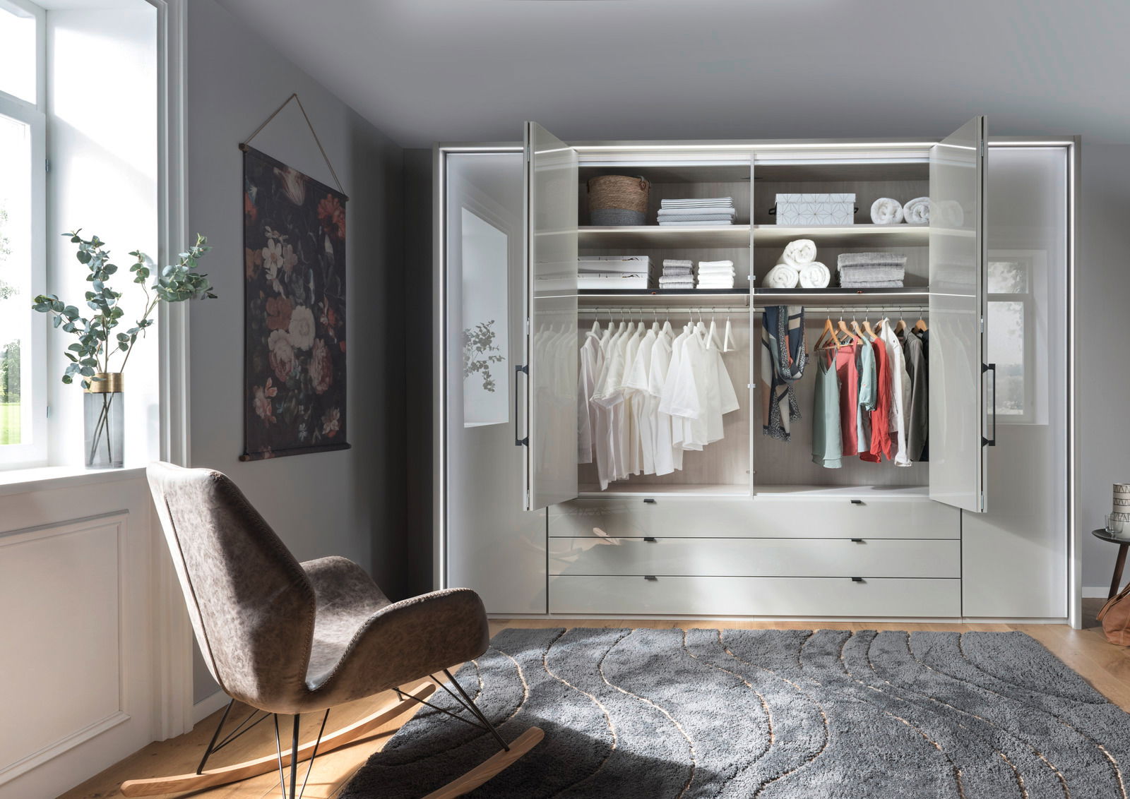 Moderner, sechstüriger Drehtürenschrank in einem stilvollen Raum, frontal fotografiert. Der Schrank ist geöffnet und zeigt ordentlich gefaltete Kleidung und Accessoires auf Regalen und Kleiderstangen.