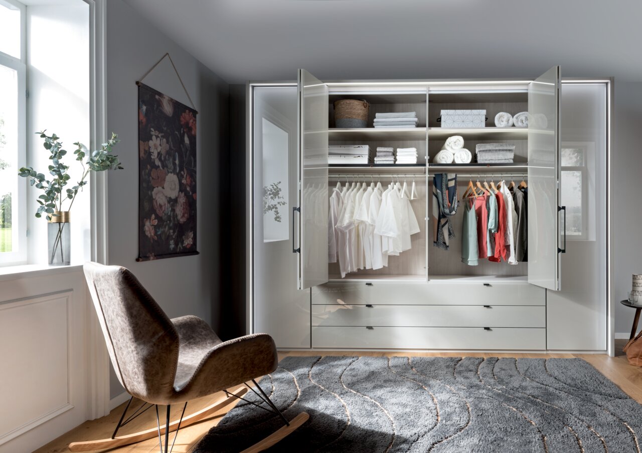 Moderner, sechstüriger Drehtürenschrank in einem stilvollen Raum, frontal fotografiert. Der Schrank ist geöffnet und zeigt ordentlich gefaltete Kleidung und Accessoires auf Regalen und Kleiderstangen.
