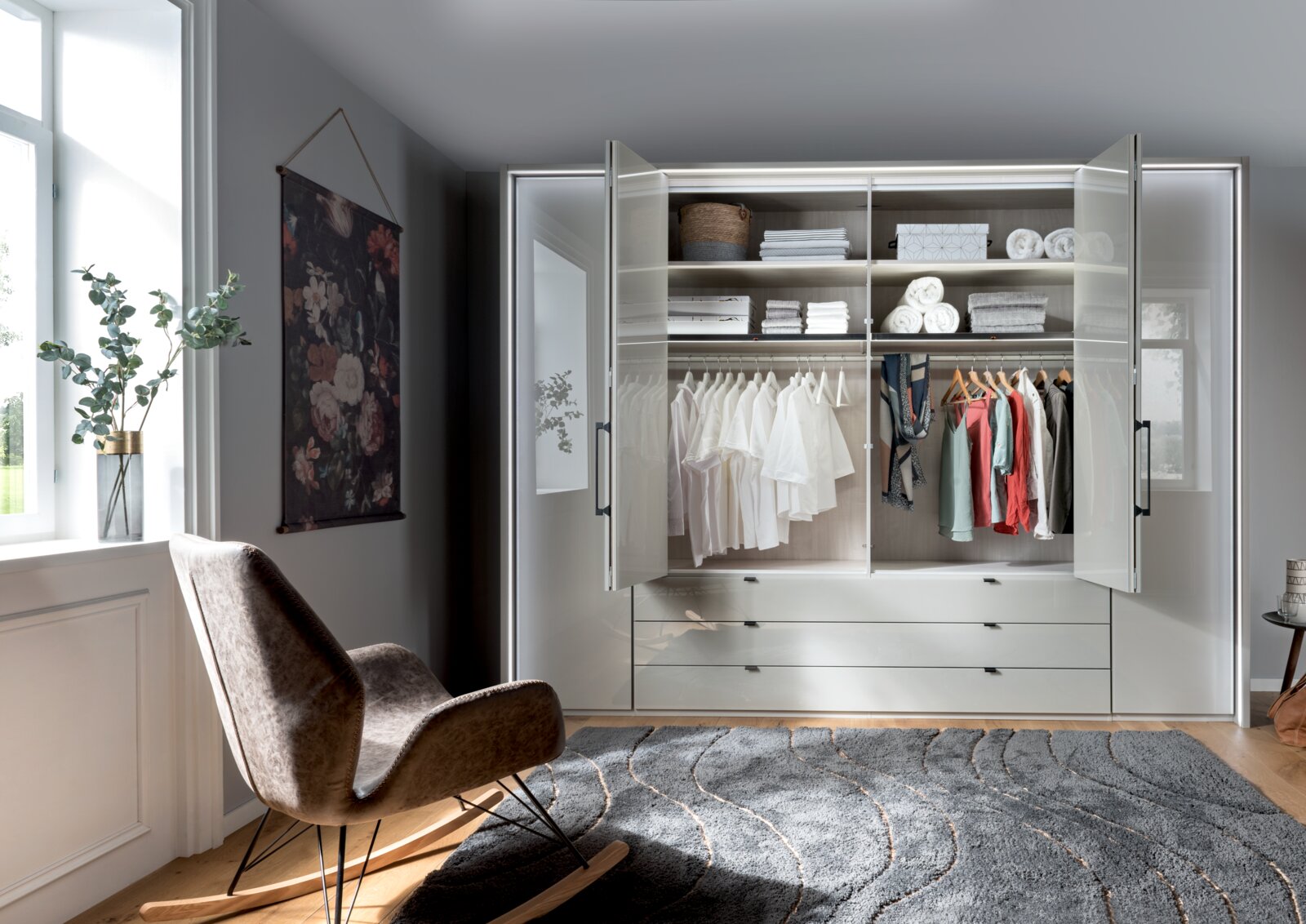 Moderner, sechstüriger Drehtürenschrank in einem stilvollen Raum, frontal fotografiert. Der Schrank ist geöffnet und zeigt ordentlich gefaltete Kleidung und Accessoires auf Regalen und Kleiderstangen.