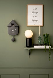 Moderne schwarze Wanduhr in Kuckucksuhren-Design, seitliche Perspektive, an grüner Wand montiert.