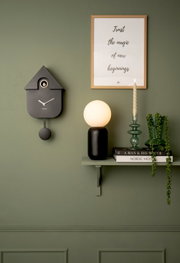 Moderne schwarze Wanduhr in Kuckucksuhren-Design, seitliche Perspektive, an grüner Wand montiert.