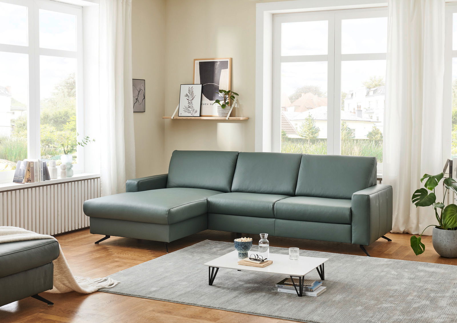 Grüne Longchairkombination in einem modernen Wohnzimmer mit großen Fenstern, Holzboden und minimalistischer Dekoration. Perspektive von vorne links.