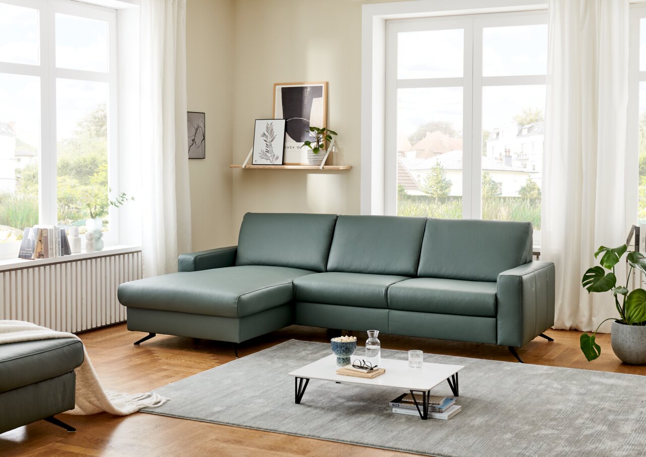 Grüne Longchairkombination in einem modernen Wohnzimmer mit großen Fenstern, Holzboden und minimalistischer Dekoration. Perspektive von vorne links.