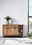 Holz-Sideboard mit zwei Schubladen und offenen Regalfächern, dekoriert mit Pflanzen und Flaschen, aus frontaler Perspektive.
