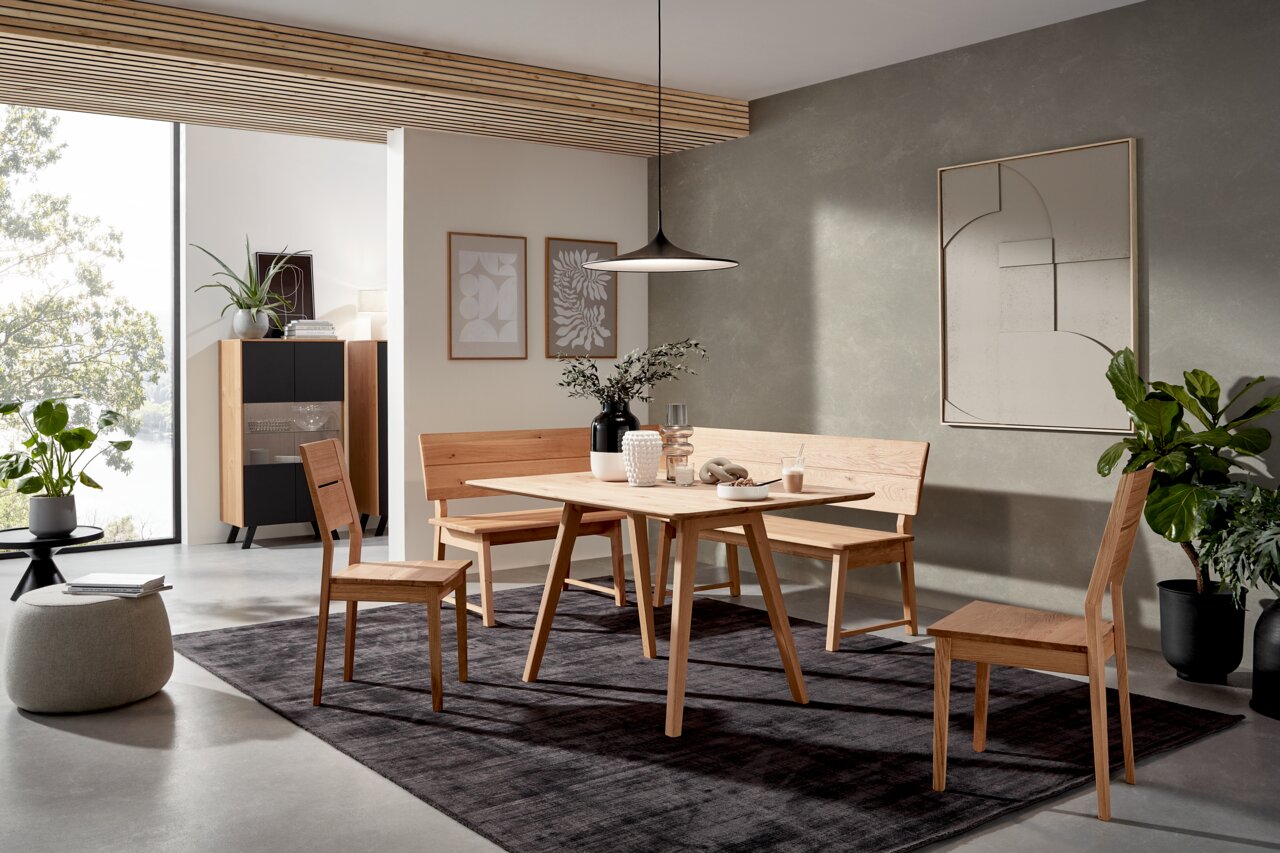Valnatura Eettafel LARINO Moderne eetkamerinrichting met houten tafel, hoekbank en stoelen in een lichte ruimte met planten en wanddecoraties, zijaanzicht.