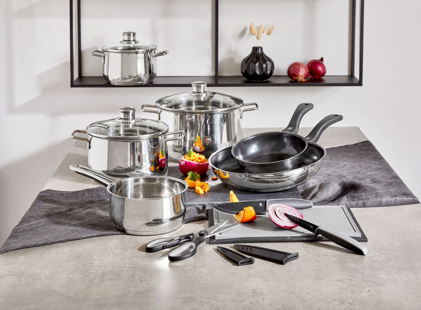 Kochtopf-Starter-Set 10-tlg. aus Edelstahl, bestehend aus mehreren Töpfen und Pfannen mit Glasdeckeln, Küchenutensilien und Schneidebrett, auf einer Küchenarbeitsplatte von vorne fotografiert.