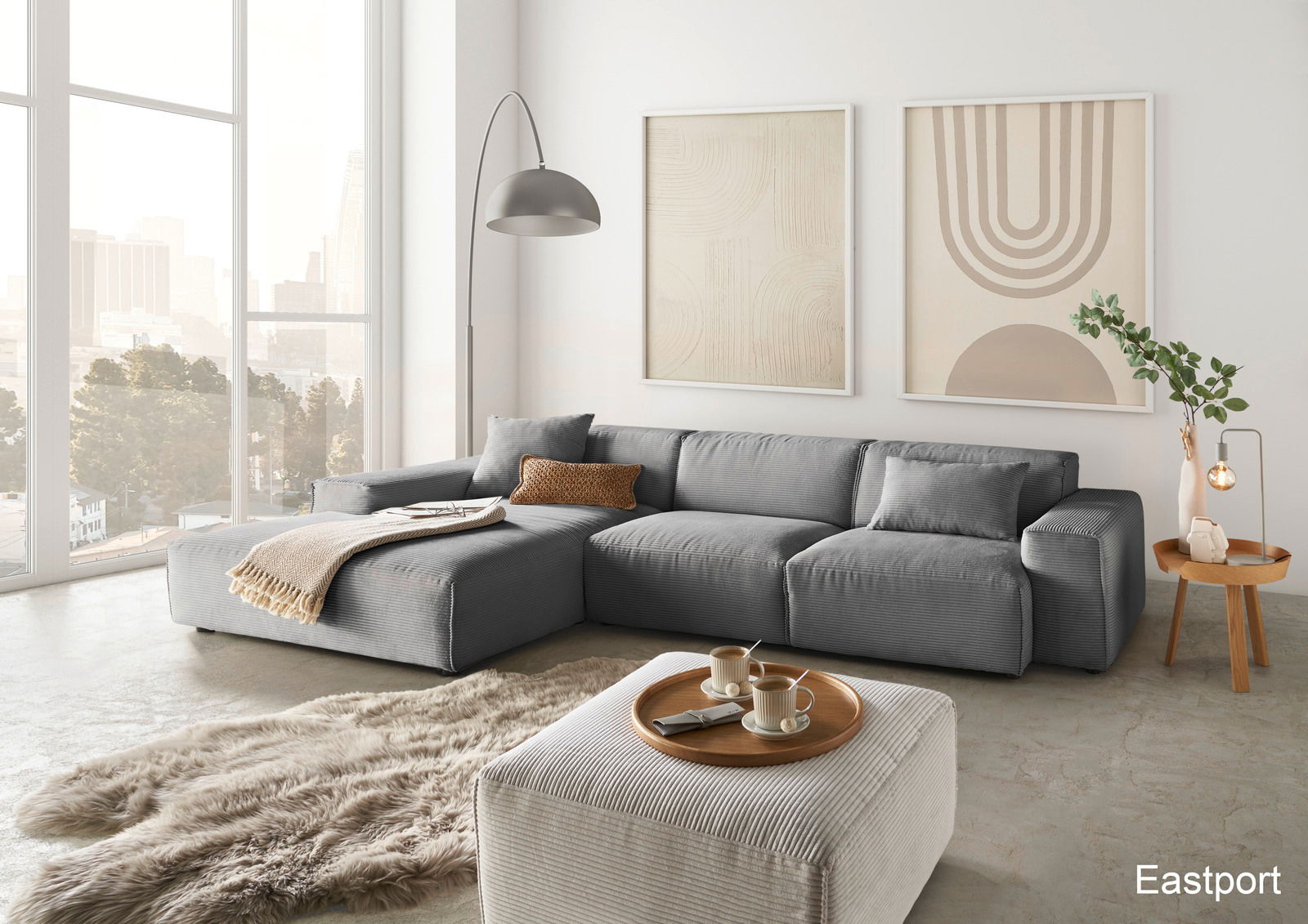 Moderne graue Rundecke mit Ottomane in einem hellen Wohnzimmer, seitliche Perspektive, dekoriert mit Kissen und Decke, umgeben von großen Fenstern und minimalistischer Kunst an der Wand.