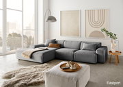 Moderne graue Rundecke mit Ottomane in einem hellen Wohnzimmer, seitliche Perspektive, dekoriert mit Kissen und Decke, umgeben von großen Fenstern und minimalistischer Kunst an der Wand.